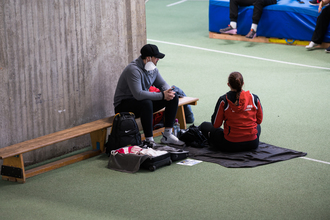 Auf dem Foto sieht man einen Sportler und eine Sportlerin in einer Trainingshalle sitzen. Der Sportler trägt eine Schutzmaske.