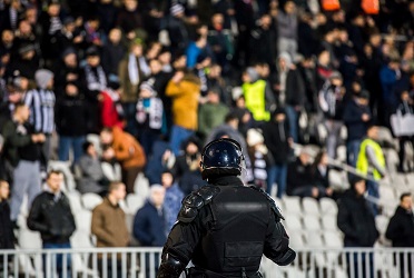 Das Bild zeigt einen Polizisten vor der mit Menschen gefüllten Tribüne eines Fußballstadions (verweist auf: BISp unterstützt BMI-geförderte Studie: Mit Stadionallianzen für sichere Fußballspiele)