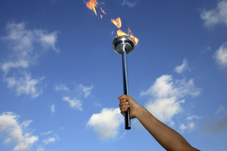 Das Bild zeigt einen Arm, der eine Olympische Fackel in die Höhe hält. Im Hintergrund ist ein blauer Himmel zu sehen.