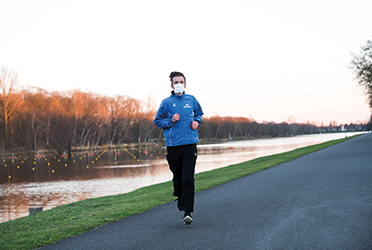 Auf dem Foto sieht man einen Jogger, der eine Atemmaske im Gesicht trägt und an einem Fluss entlang läuft. (verweist auf: Ausdauerleistung und Wohlbefinden von Athletinnen und Athleten in Zeiten von COVID-19)