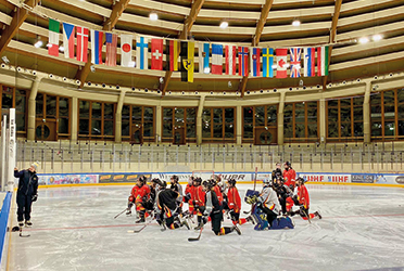 Eishockeyspielerinnen knien auf einer Eisfläche und schauen zu einer Trainerin. Diese zeigt etwas auf einer Tafel.