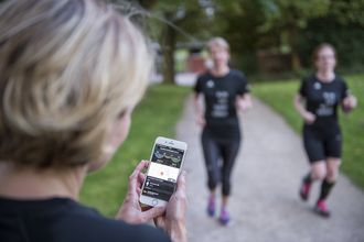 Auf dem Bild sieht man eine Frau von hinten, die ein Smartphone mit einer App darauf in der Hand hält. Im Hintergrund joggen zwei Frauen.