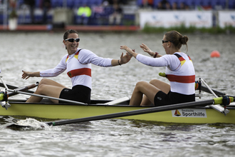 Das Bild zeigt zwei Sportlerinnen in einem Ruderboot auf dem Wasser, die sich abklatschen.